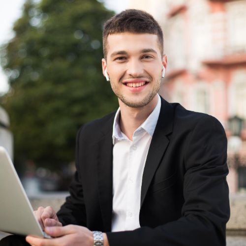 young cheerful man classic black suit white shirt with wireless earphones happily lookingin camera with laptop hands while spending time outdoor (1)