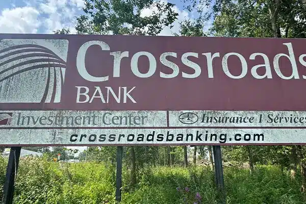Faded maroon roadside sign reading Crossroads BANK and crossroadsbanking.com, above a grassy verge with trees in the background.