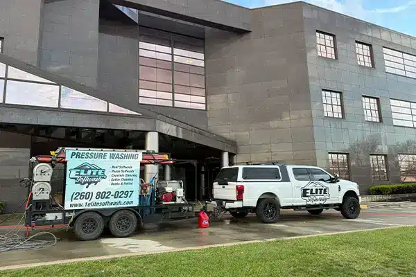 White pickup truck with Elite Softwash logo towing a pressure-washing equipment trailer parked outside a multi-story office building with large windows