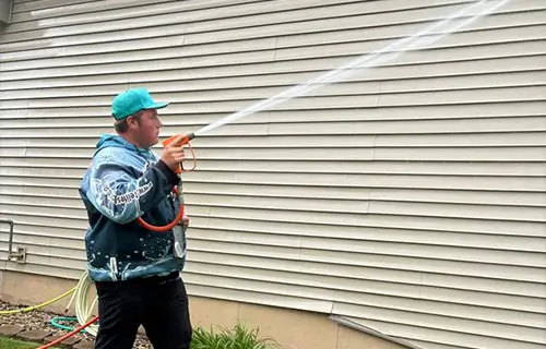 Person wearing teal cap and patterned jacket spraying a strong stream of water from a hose at house vinyl siding.