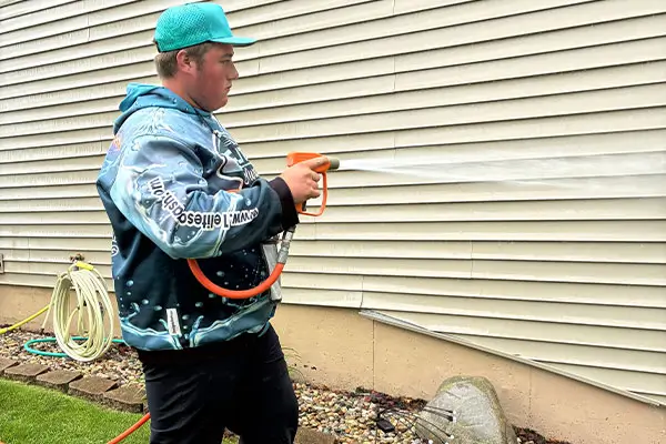 Person in teal cap and patterned blue hoodie pressure-washing beige vinyl house siding with an orange hose and nozzle, water spraying.