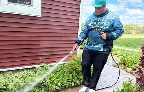 Person in teal cap and blue hoodie spraying water from a hose onto plants and a stone path beside red house siding