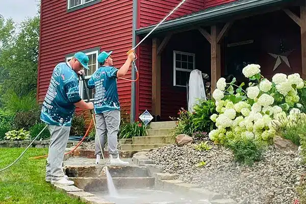 Two workers in matching shirts pressure-washing stone steps of a red-sided house near white hydrangeas.