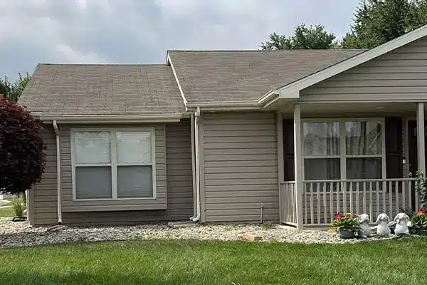 Single-story beige siding house front with small porch and railing, two windows, green lawn, gravel border, potted flowers and three garden statues.