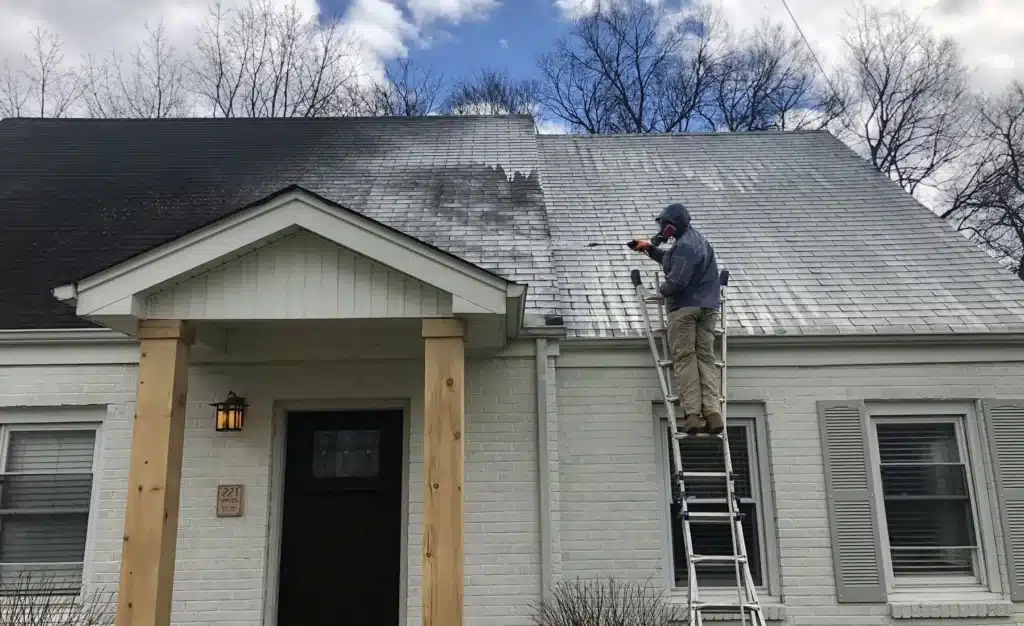 Person on ladder pressure-washing a gray shingle roof, revealing a lighter cleaned area above a white brick house.